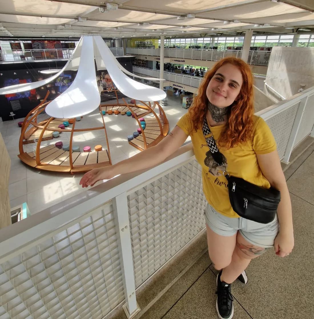 A Gabi sorrindo numa passarela de biblioteca, de camiseta amarela, com uma estante circular de madeira ao fundo